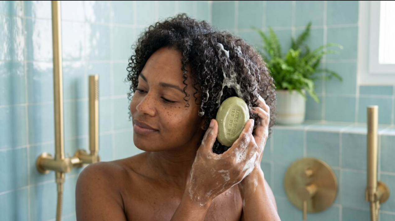 femme aux cheveux boucles utilisant un shampoing solide sous la douche